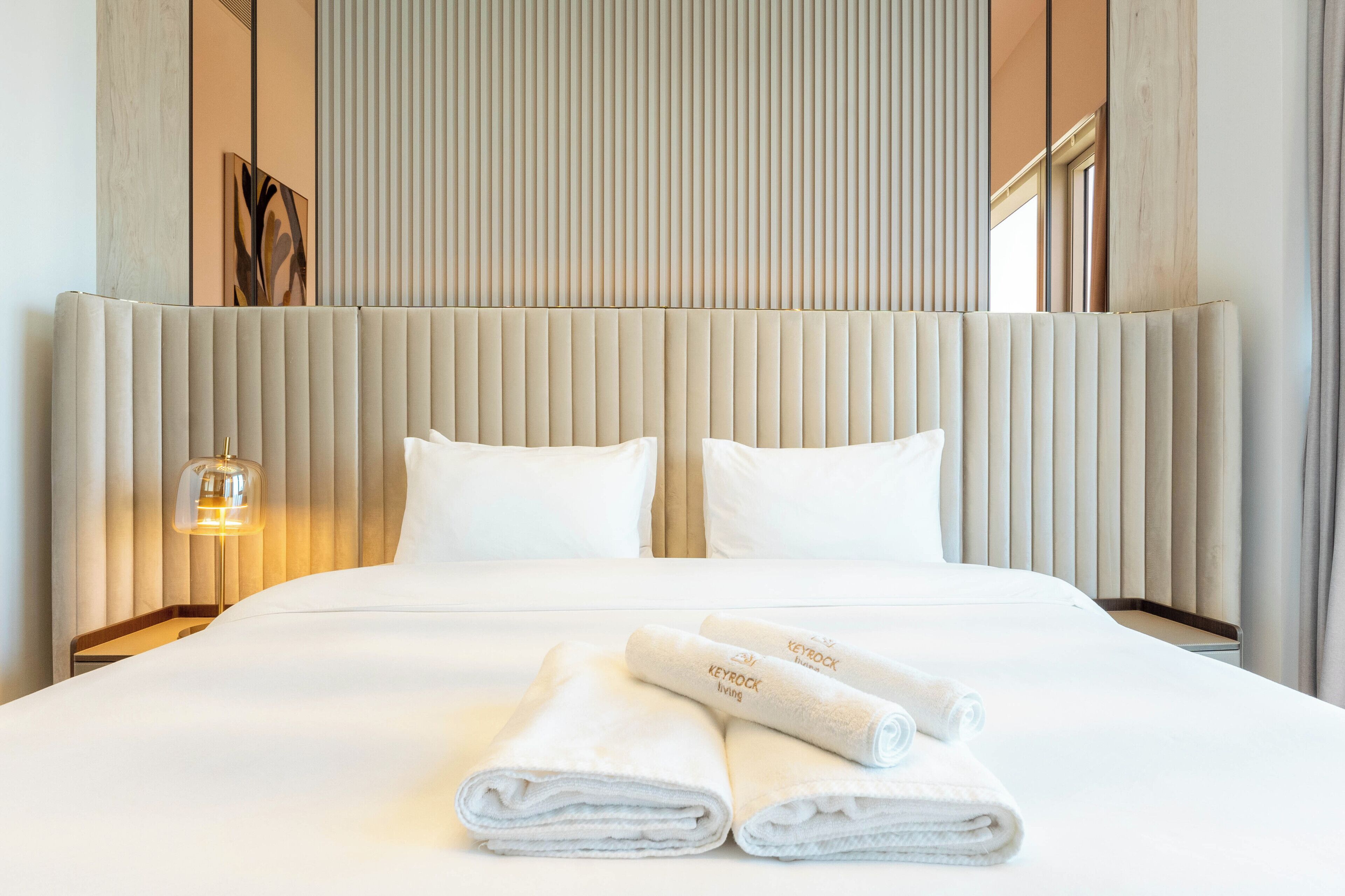 A hotel room with a large bed, two pillows, a bedside lamp, and rolled towels on the bed.