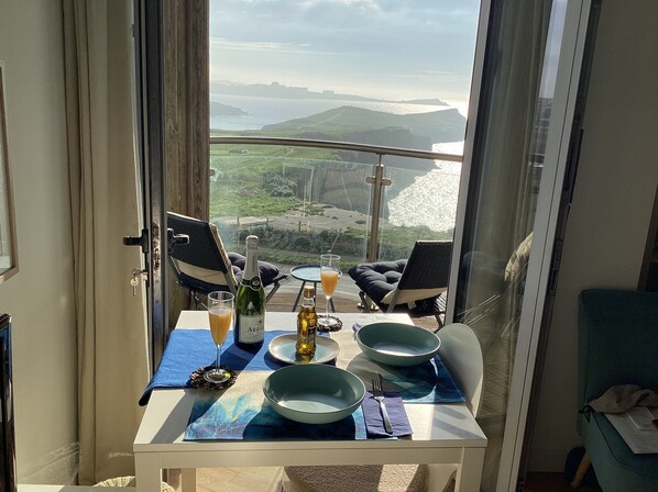 A balcony table with chairs, a bottle of wine, glasses, and a view of the coastline. - Ocean Front top floor apartment, with far-reaching views. Sleeps 4. (Newquay)