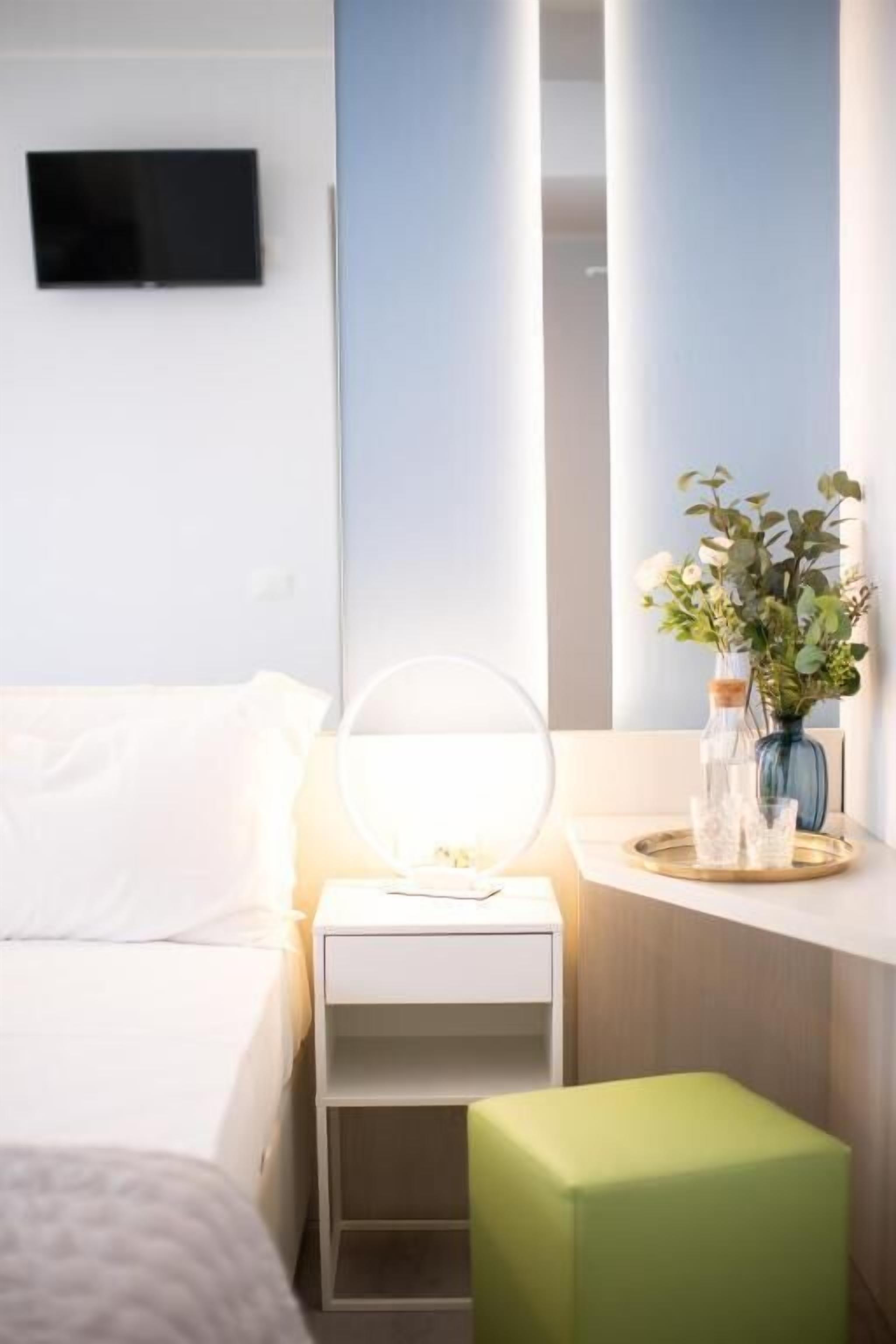 A hotel room with a bed, a bedside table with a lamp, a mirror, a small stool, and a vase with flowers.