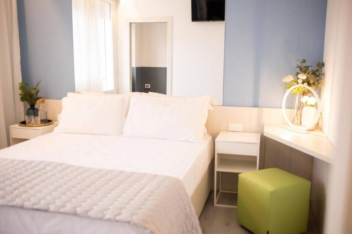 Una habitación de hotel con una cama, mesitas de noche, un reposapiés verde, un espejo y un televisor en la pared.