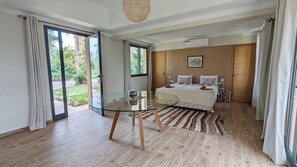 Ein Schlafzimmer mit Glastisch, Bett mit Kissen, Holzboden und Blick ins Grüne.