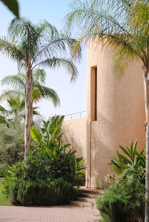Ein Gebäude mit einer beige farbenen Fassade, einer Treppe zum Eingang und umgebendem Grün, darunter Palmen und Sträucher.