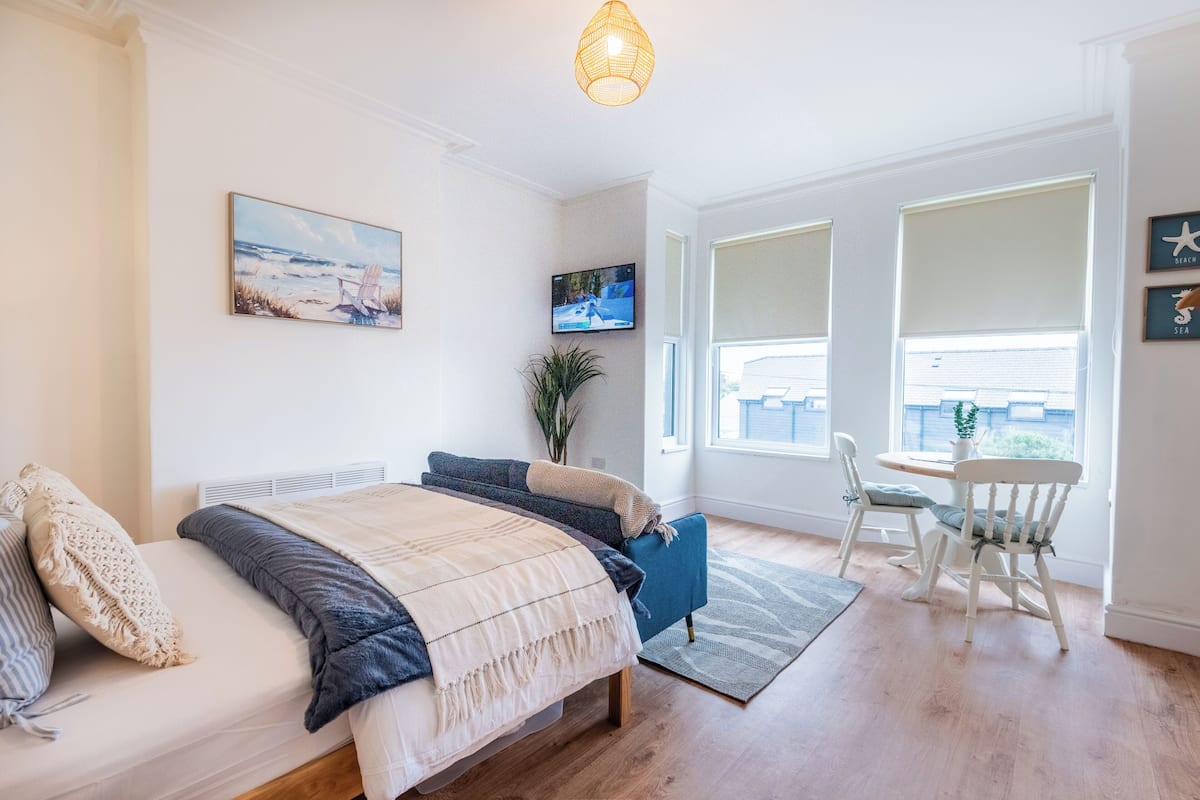 Un dormitorio con cama, televisor, planta y zona de comedor con sillas.