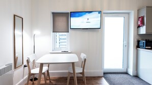 Una habitación moderna con una mesa blanca, dos sillas, un televisor montado en la pared, una ventana con persianas y una puerta.