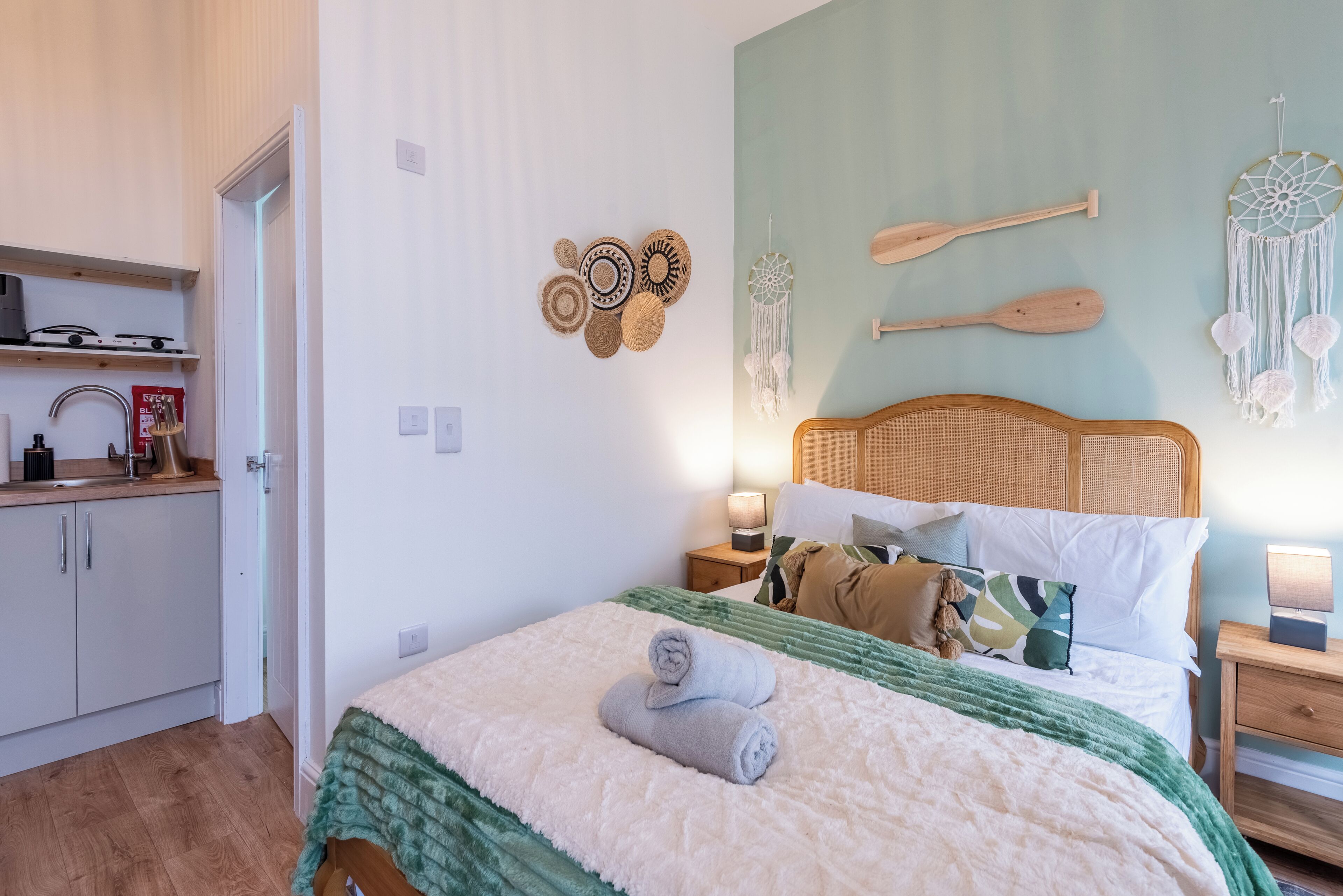 A bedroom with a bed, pillows, a bedside table, a nightstand, a wall with decorative items, and a kitchen area with cabinets and a stove.