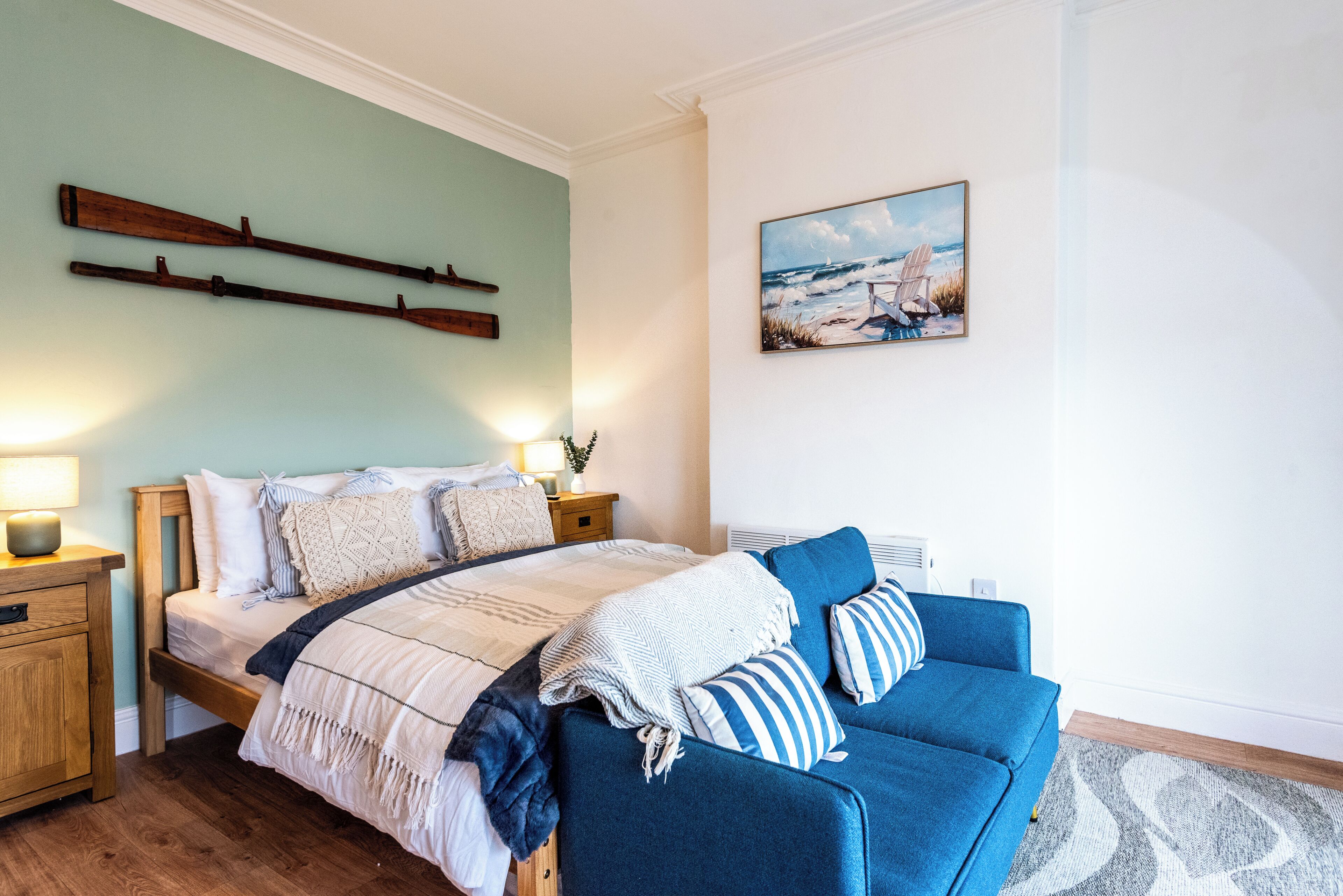 A bedroom with a bed, a blue sofa, wooden headboard, bedside tables, a painting, and a wall-mounted shelf.