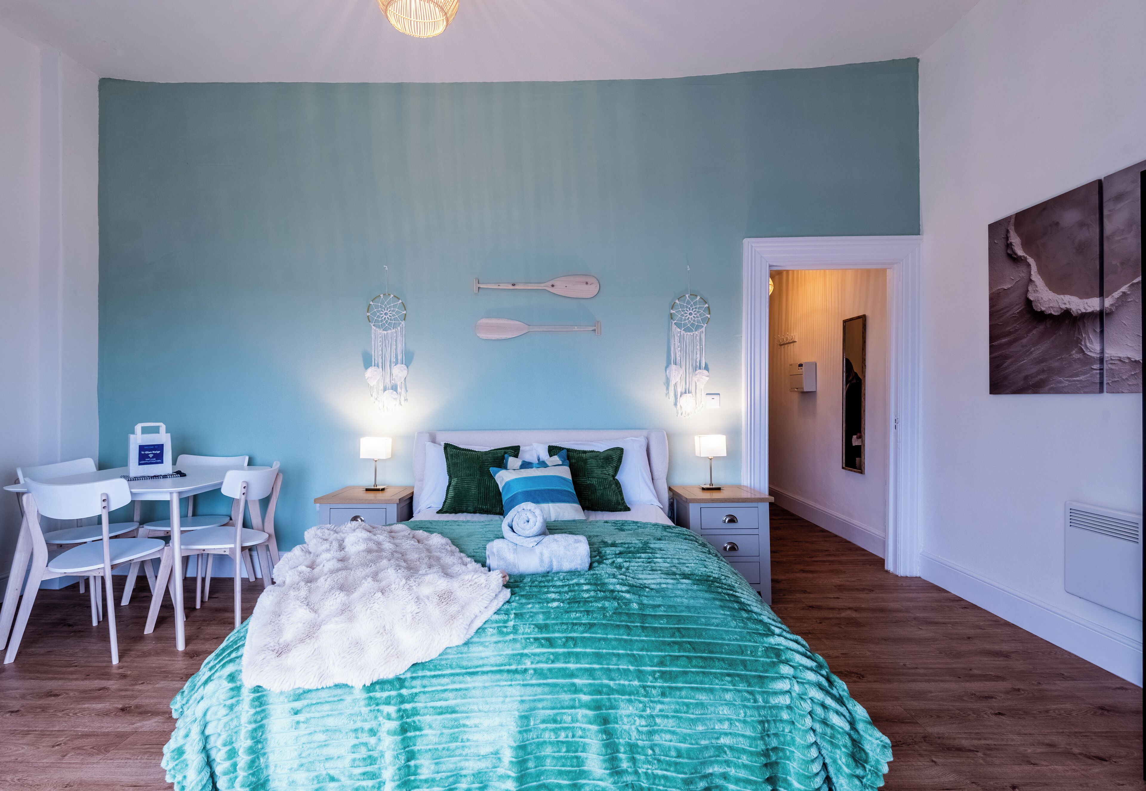 Un dormitorio con cama, mesitas de noche, escritorio, sillas y una pared decorada con atrapasueños.