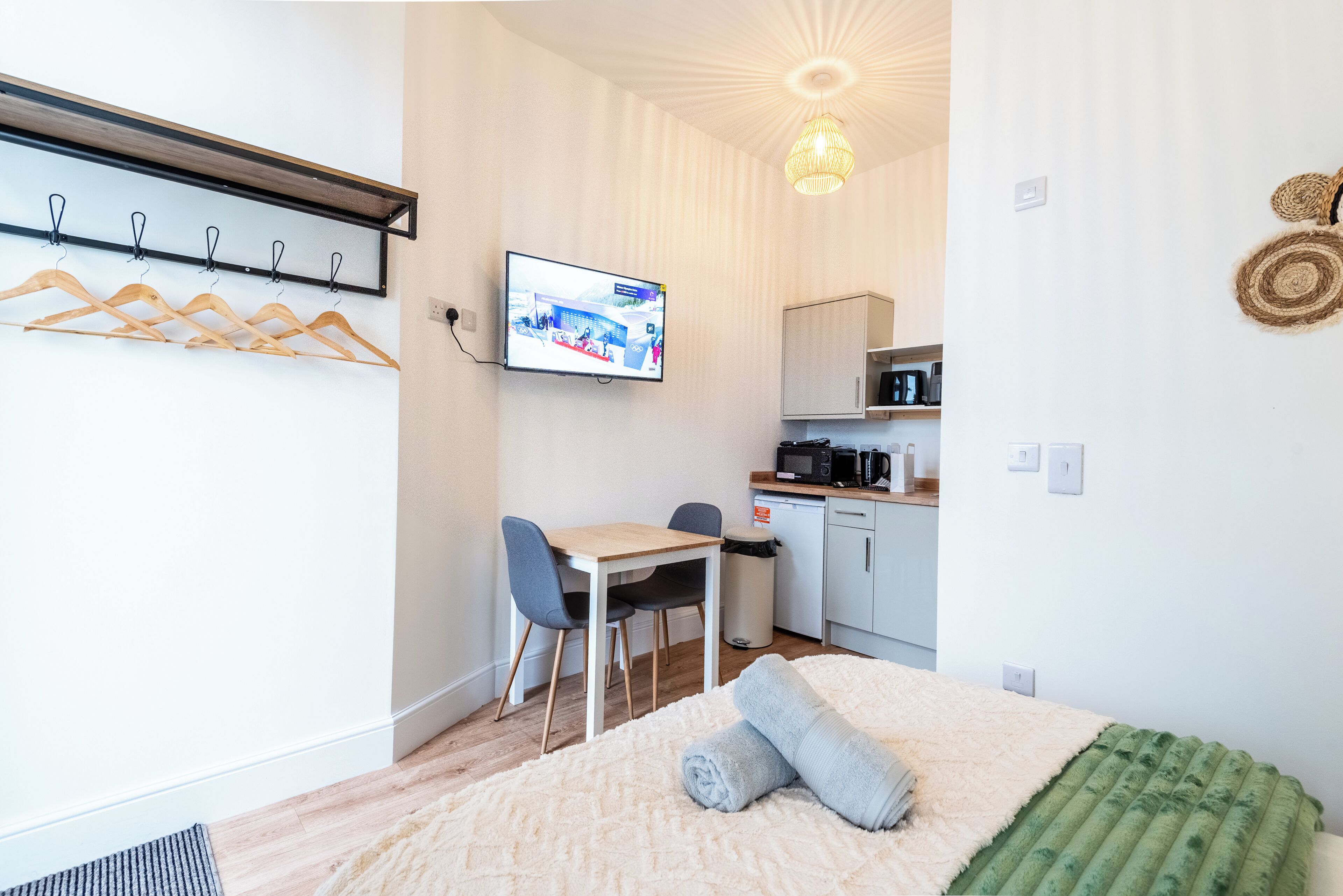 A room with a bed, a desk, chairs, a television, a kitchen area, and a coat rack.