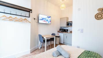 A room with a bed, a desk, chairs, a television, a kitchen area, and a coat rack.