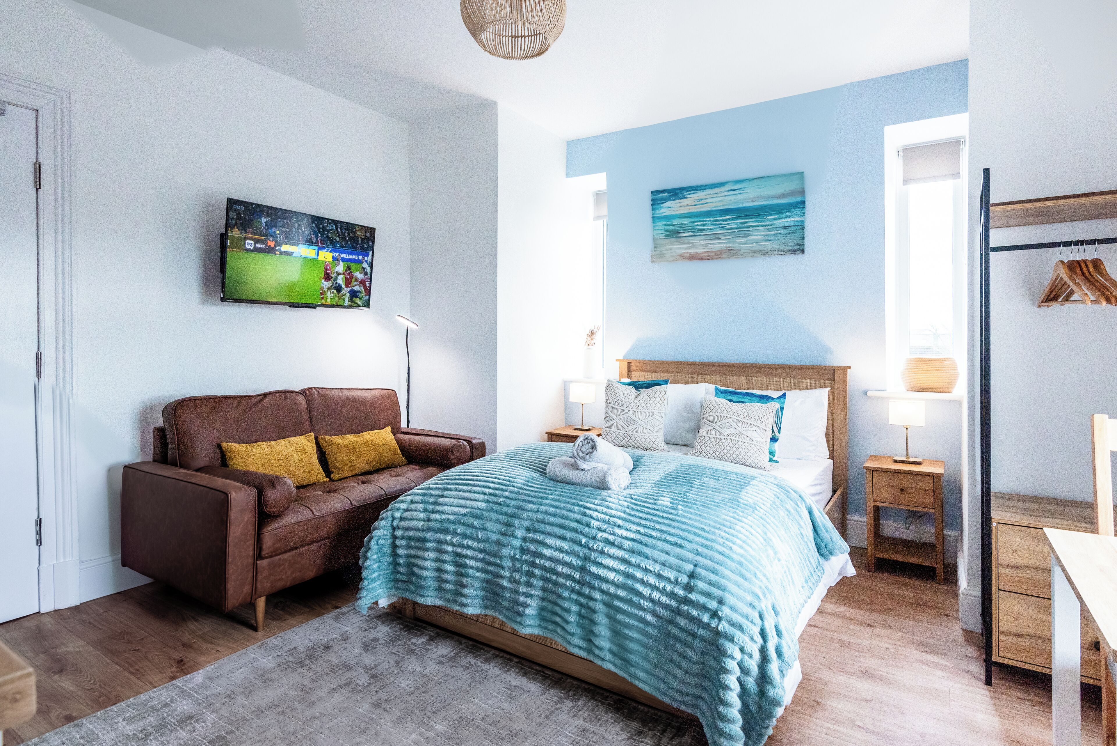 A bedroom with a bed, a brown leather sofa, a television on the wall, and a painting.