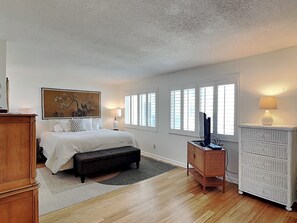 A bedroom with a bed, bedside lamps, a dresser, a television, and a window with blinds. - Bent Palm 302 | Romantic Condo + Remodeled Touches (Ormond Beach)