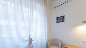 Een kamer met een raam met dunne gordijnen, een stoel, een klein houten kastje en een wandairconditioner.