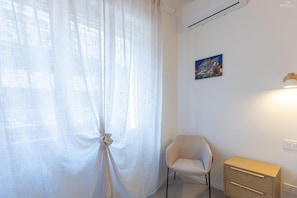 A room with a window covered by sheer curtains, a chair, a small wooden cabinet, and a wall-mounted air conditioner. - Sivori Apartment (Rome)