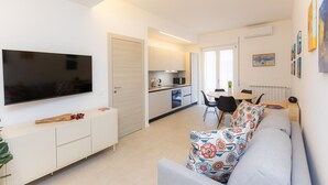 Een moderne woonkamer met een flatscreen televisie, een bank met bloemenkussens, een eethoek met tafel en stoelen, en een keuken met kastjes en een magnetron.