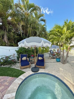 En baghave med swimmingpool, to blå liggestole, en hvid parasol og palmer.