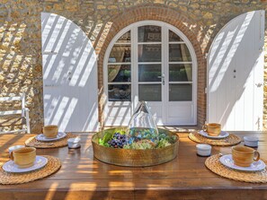 Une table dressée avec un centre de table composé de fruits et un vase en verre, des portes blanches et un mur en pierre.