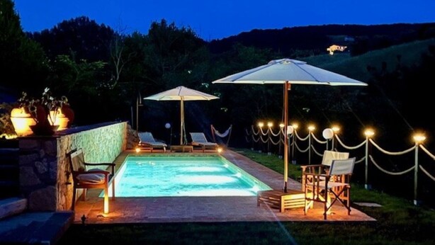 Una piscina con sillas de descanso y sombrillas, rodeada por un muro de piedra y una valla.