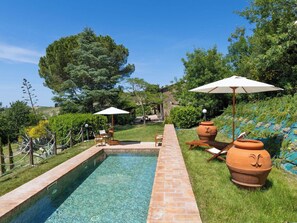 Ein Schwimmbecken mit Ziegelkanten, umgeben von Gras und Bäumen, mit Sitzgelegenheiten und Sonnenschirmen im Freien.