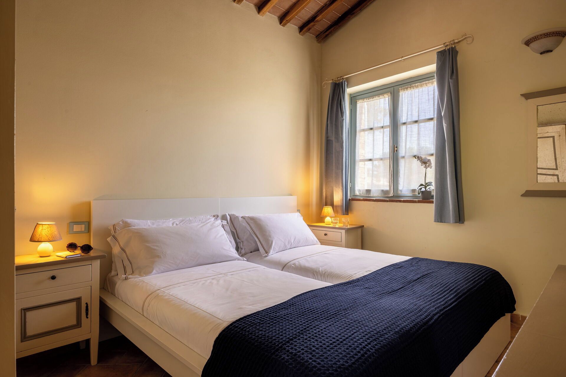 A bedroom with a bed, bedside lamps, a window with curtains, and a small potted plant.