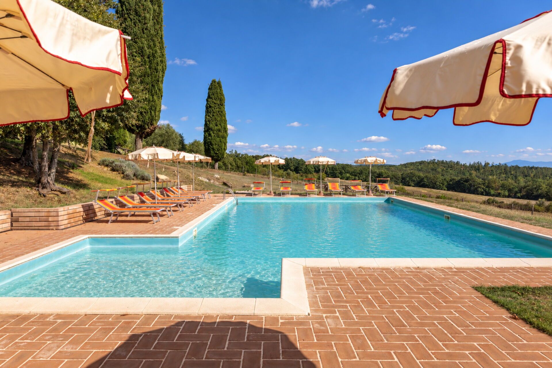 A swimming pool with lounge chairs and umbrellas, surrounded by a brick patio and a view of a hilly landscape.