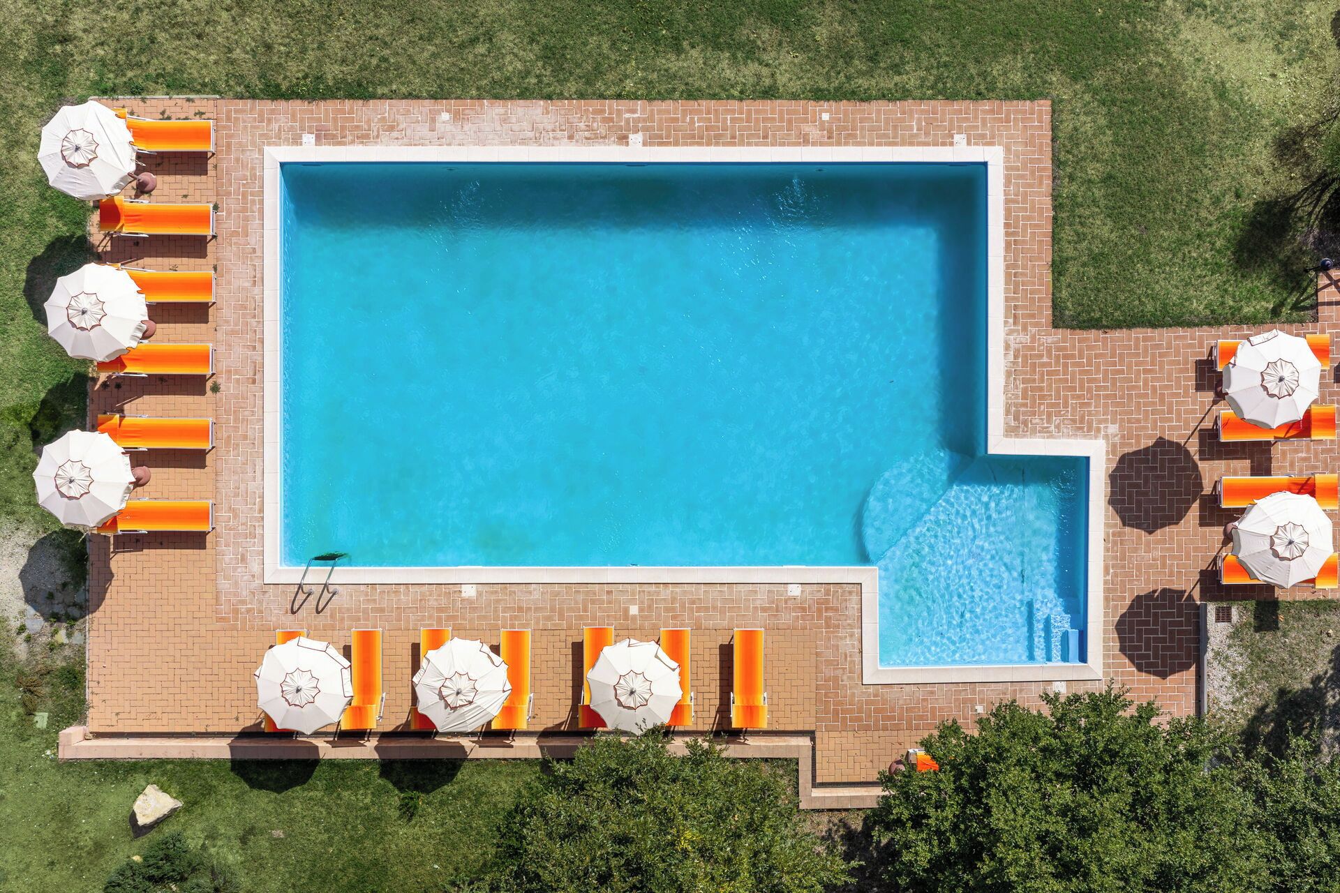 Eine Vogelperspektive auf einen rechteckigen Swimmingpool mit Ziegelumrandung, umgeben von orangefarbenen Liegestühlen und weißen Sonnenschirmen.