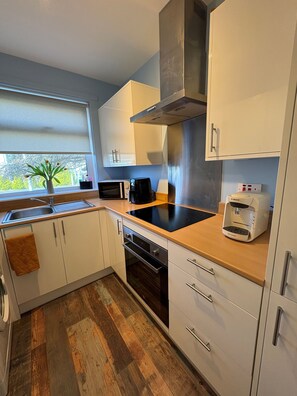 A modern kitchen with white cabinets, a black induction cooktop, a Bosch coffee machine, and a window with a view of trees. - Stylish 2 bedroom Cottage Near Stirling + Parking & Pets (Stirling)