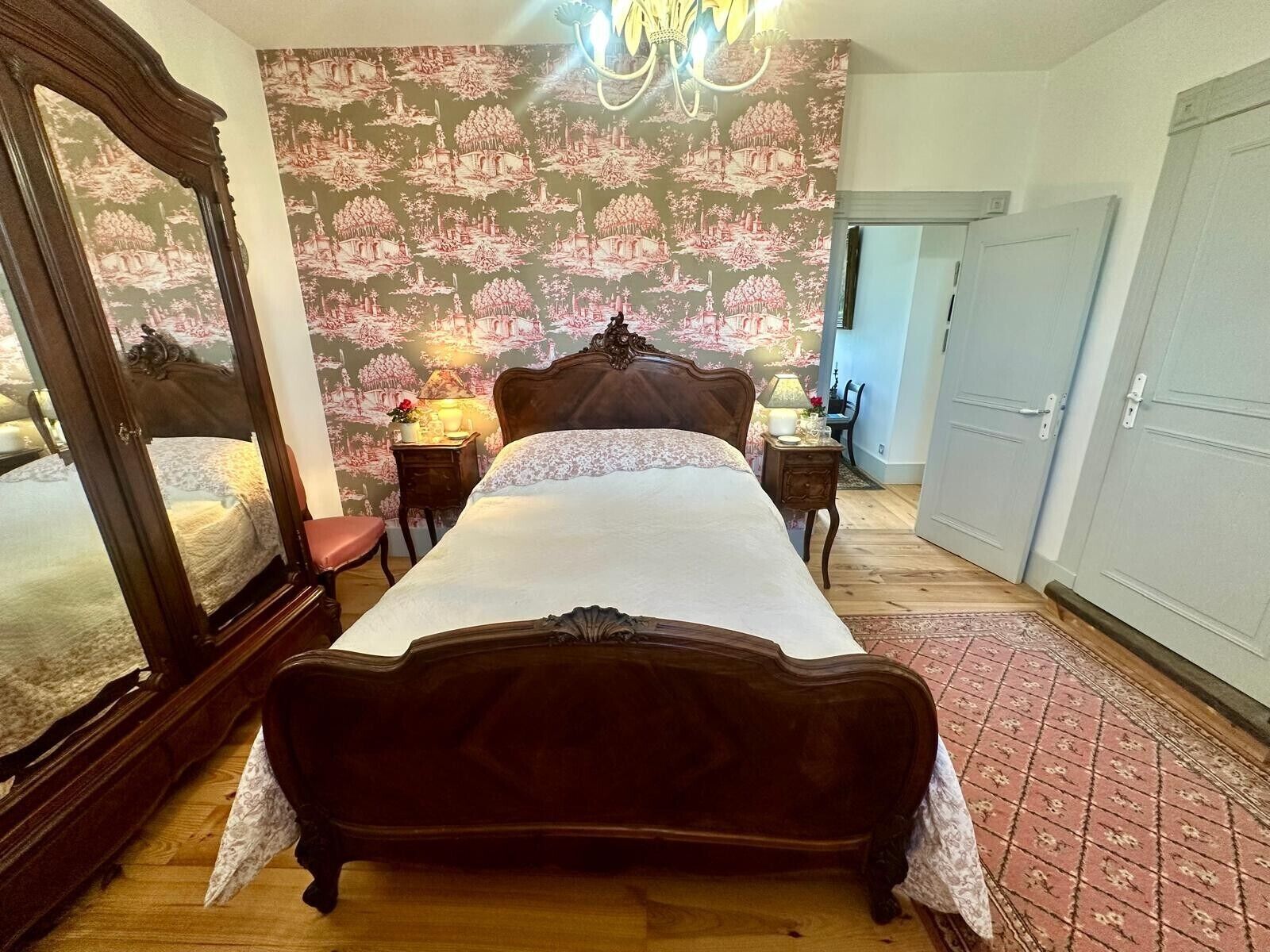 A bedroom with a large bed, a wooden wardrobe, a nightstand with a lamp, and a patterned wallpaper.