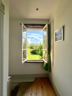 Une pièce avec une fenêtre donnant sur un jardin, un tableau encadré au mur et un plancher en bois.