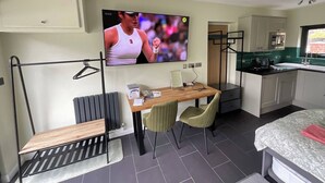Habitación compacta con televisor, una pequeña mesa de comedor, dos sillas, una banca, radiador, zona de cocina con armarios y una ventana.