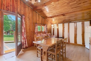 Un intérieur en bois avec une table à manger, des chaises et une vue sur un paysage verdoyant, visible à travers une porte.