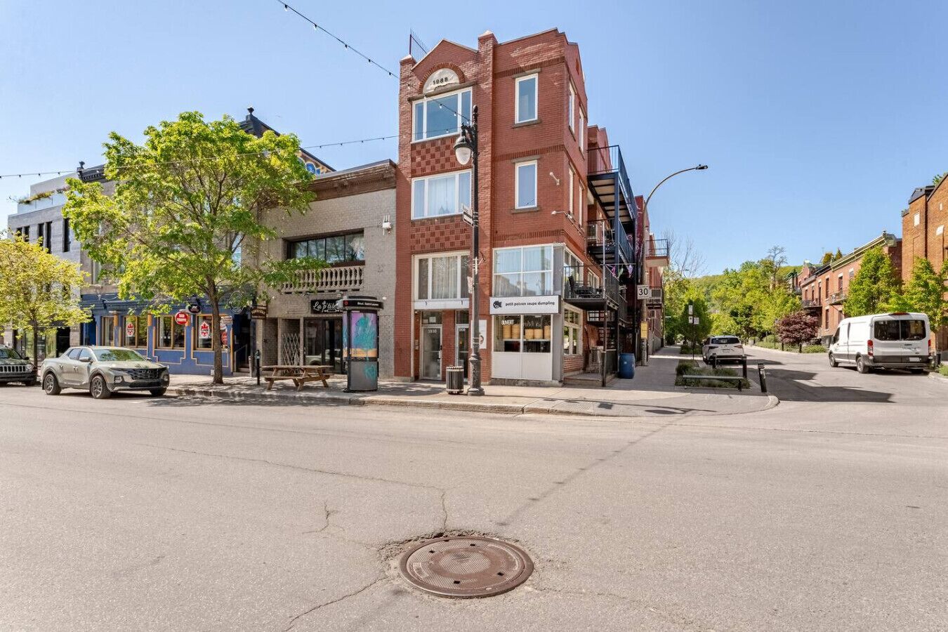 A street view with a brick building, a sidewalk, a manhole cover, and parked cars.