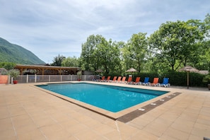 Une piscine rectangulaire, bordée d’une terrasse carrelée, entourée de chaises longues et de parasols, le tout sur fond de verdure, avec des arbres et une montagne en arrière-plan.