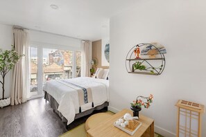 Ein Schlafzimmer mit Bett, einem Fenster mit Vorhängen, Blick auf Gebäude, einer Wandregal mit Dekoration und einem kleinen Tisch mit Teekanne und Tassen.