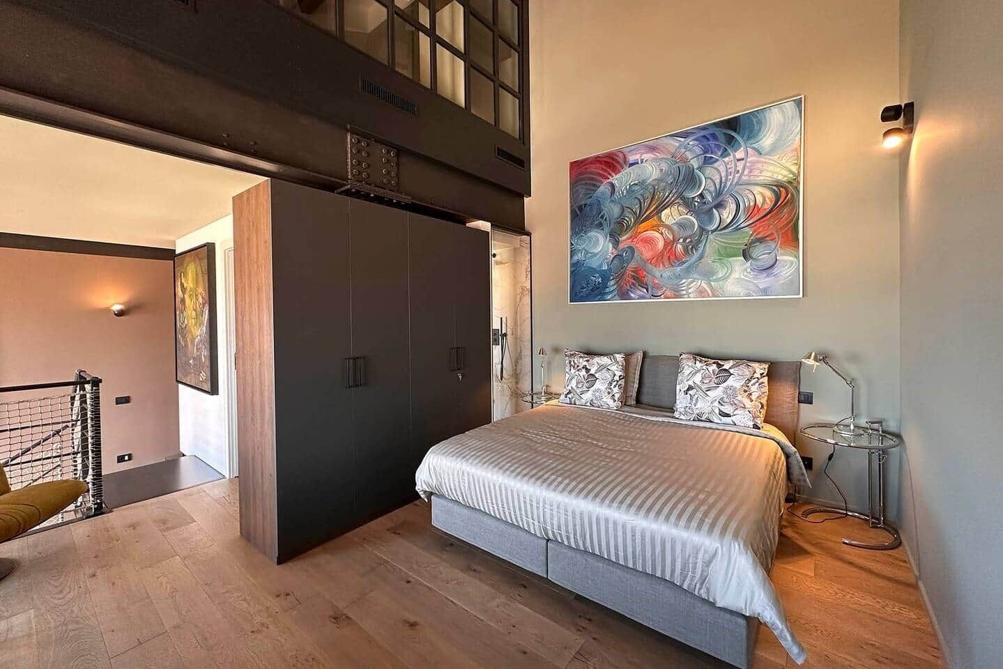 Ein Schlafzimmer mit einem Bett, einem großen abstrakten Gemälde, einem Schreibtisch mit Lampe und einer Treppe, die zu einer oberen Ebene führt.