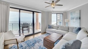 Ein modernes Wohnzimmer mit einem weißen Sofa, einem Holztisch, einem Deckenventilator und Meerblick durch große Fenster.