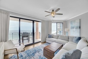 Ein modernes Wohnzimmer mit einem weißen Sofa, einem Holztisch, einem Deckenventilator und Meerblick durch große Fenster.