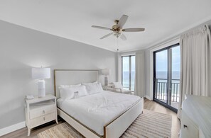Ein Hotelzimmer mit einem großen Bett, einem Deckenventilator, einem Nachttisch mit Lampe, Blick auf den Ozean durch eine Balkontür und einem Fenster mit Vorhängen.