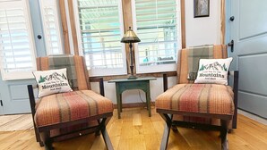Una habitación con dos sillones, una mesita y una lámpara. Hay ventanas con persianas, una puerta y un cuadro enmarcado en la pared.