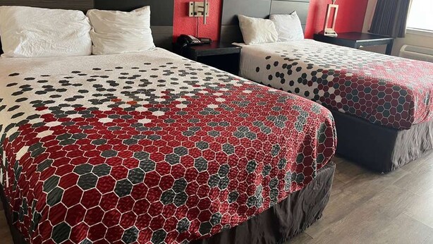 Two beds with patterned bedding, a red accent wall, a bedside table with a lamp, and a window with curtains. - FairBridge Inn near Riverwalk - Alamo (San Antonio)
