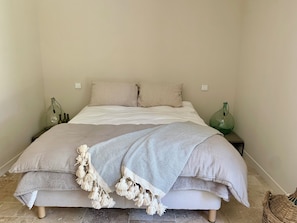 Ein Bett mit einer beigen Decke und Kissen, zwei Nachttischlampen und ein gewebter Korb am Boden.