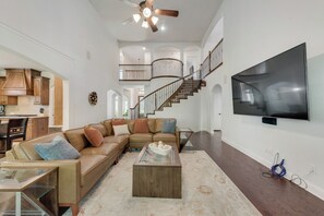 A spacious living area with a large sectional sofa, a coffee table, a TV mounted on the wall, and a staircase leading to an upper level. - The Full Package (Grand Prairie)