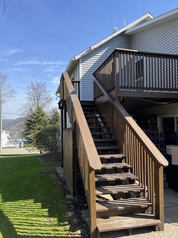 Un escalier en bois mène à un balcon d’où l’on peut admirer une cour verdoyante et une maison en arrière-plan.