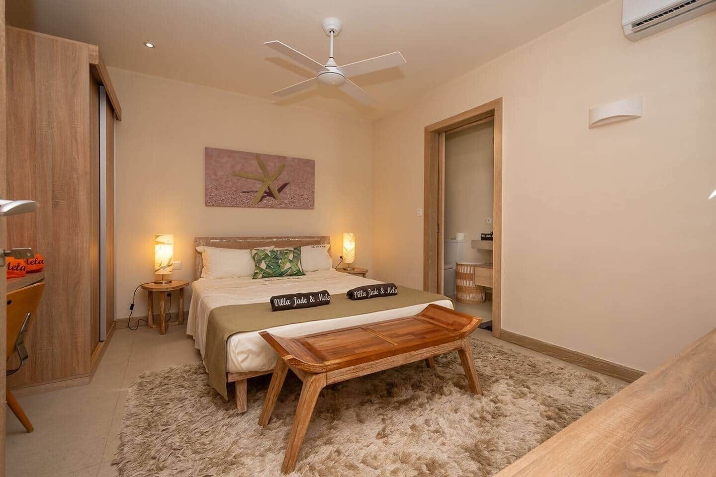 A bedroom with a bed, bedside lamps, a ceiling fan, a wooden bench, and a painting on the wall.