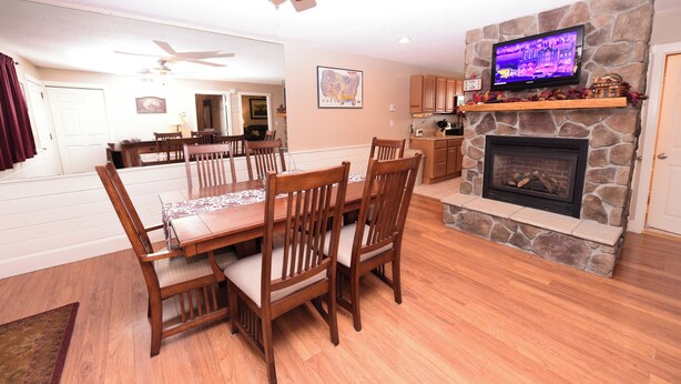 A dining area with wooden chairs and a table, a stone fireplace, a television mounted on the wall, and a ceiling fan. - Steps to Settlers Green | 3BR, 2 Bath, Sleeps 8 | Central AC + Fireplace (North Conway)