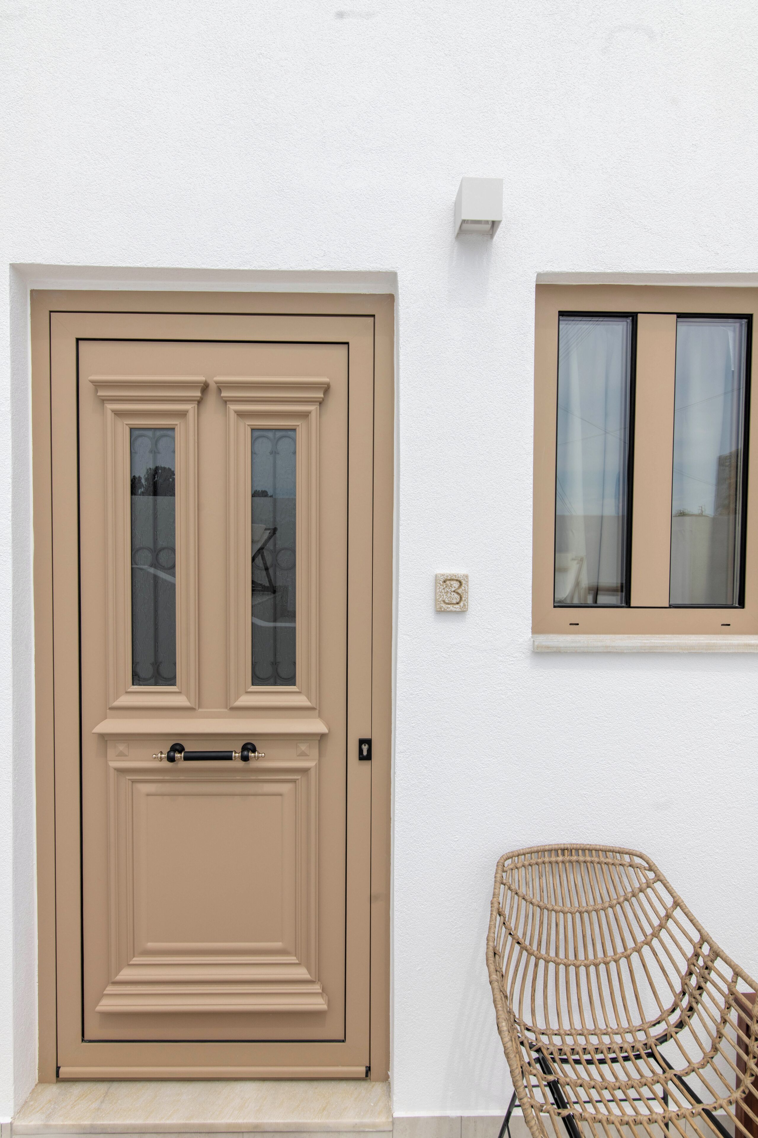 Una porta beig amb un pom, una finestra amb cortines i una cadira de vimet.