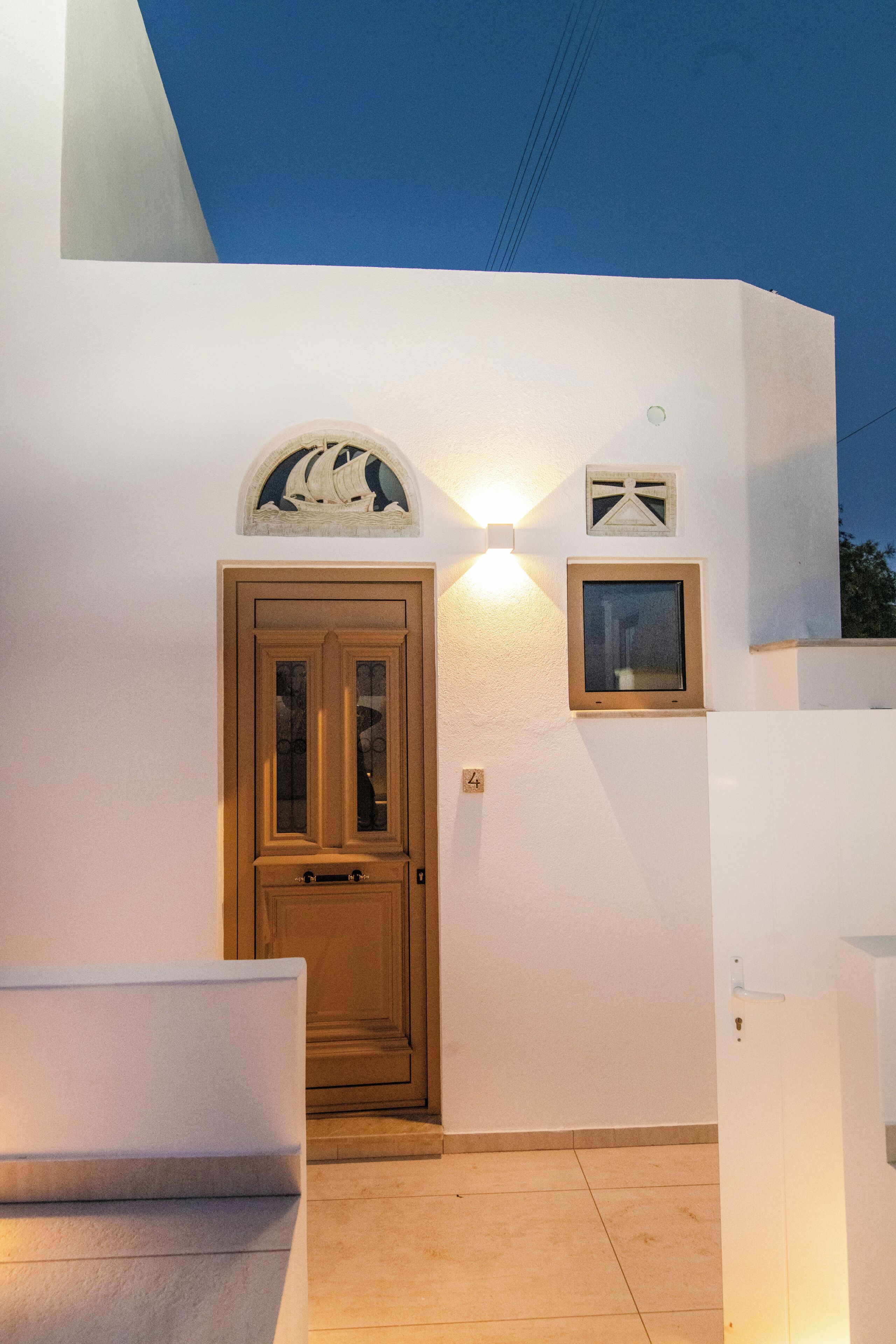 A white building with a wooden door, a small window, and a wall-mounted light fixture.