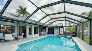 Ein überdachter Pool mit Glasdach, angrenzende Liegestühle und Blick auf Palmen.