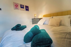 A neatly made bed with green towels, a bedside lamp, and two wall art pieces depicting beach and Hawaii themes. - Stylish 2bdr 10m to City Centre Free Parking (Manchester)