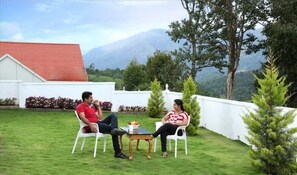 A garden with a white building, a table with fruit, and a mountain in the background. - THE FOG Munnar Resorts AND SPA (Devikolam)