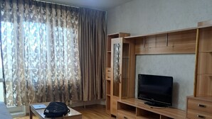 Ein Wohnzimmer mit einer Couch, einem Fernseher auf einem Holzschrank, einem Tisch mit einem Laptop und einer Tasche, sowie einem gemusterten Vorhang.